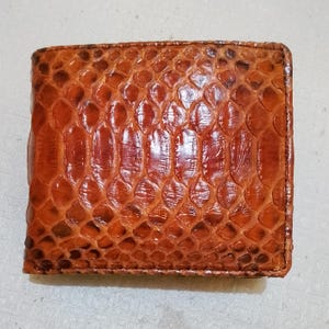 May include: A rectangular bifold wallet featuring a glossy, textured surface with a distinct brown and reddish-brown scale pattern. The exterior displays a repeating, overlapping scale design that covers the entire front face of the accessory.