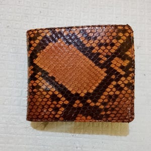 Könnte beinhalten: Ein Klappgeldbörse mit einem strukturierten Schlangenhautmuster in Braun- und Dunkelbrauntönen. Die Außenseite zeigt ein sich wiederholendes schuppenartiges Design mit einer ausgeprägten geometrischen Anordnung dunkelbrauner Markierungen.