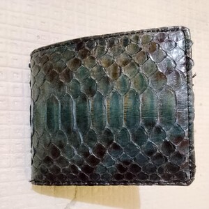 May include: A bi-fold wallet featuring a textured snakeskin pattern in deep shades of teal, dark green, and black. The surface displays a repeating scale design with a glossy finish, showcasing the natural organic texture of the material.