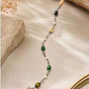 May include: A delicate bracelet featuring a silver-toned metal chain interspersed with a variety of beads in shades of green, white, and yellow, finished with a light-colored wooden bead segment at one end and a circular white button-style clasp.
