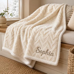 Könnte beinhalten: Eine cremefarbene Plüschdecke mit strukturiertem Fischgrätenmuster und weichem Fleece-Rand. Der Name Sophia ist in einer geschwungenen Schreibschrift mit hellbraunem Garn in die untere Ecke der Decke gestickt.