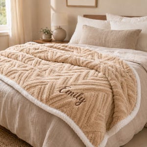 May include: A tan, textured throw blanket with a raised herringbone pattern and a white sherpa-style border. The name Emily is embroidered in a dark brown script font on the lower right corner of the blanket, which is draped over a bed.