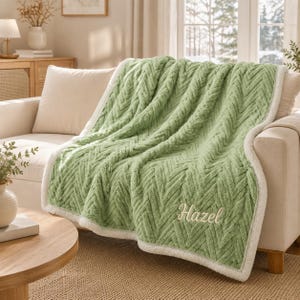 Könnte beinhalten: Eine salbeigrüne Wohndecke mit strukturiertem Fischgrätenmuster und einem weißen Sherpa-Fleece-Rand. Die Decke ist mit dem Namen Hazel in Schreibschrift in der unteren rechten Ecke bestickt und über ein helles Sofa drapiert.