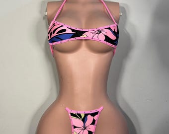 May include: A two-piece bikini set featuring a black base with a vibrant pink, blue, and yellow floral pattern. The top has a halter neck design and the bottom is a high-cut thong style. Both pieces are trimmed with bright pink fabric and small studs.