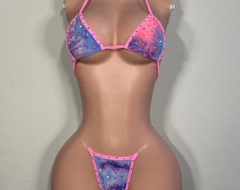Può includere: Un set bikini a due pezzi con una stampa ispirata alla galassia nei toni del blu, viola e rosa. Il top a triangolo e lo slip a perizoma sono rifiniti con tessuto rosa acceso e decorati con strass trasparenti sparsi su tutto il capo.