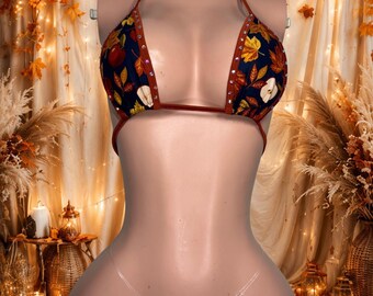 Può includere: Un set bikini a due pezzi in tessuto blu navy scuro con un motivo ripetuto di foglie autunnali e fette di mela. Il top a triangolo e lo slip sono bordati con tessuto color ruggine e decorati con piccoli strass trasparenti e scintillanti.