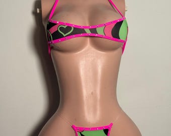 Può includere: Un set bikini a due pezzi con un vivace motivo di cuori e forme floreali neri, verde lime e rosa acceso. Il top è in stile allacciato al collo con bordi rosa acceso e piccoli strass incastonati. Lo slip è un perizoma con spalline trasparenti.
