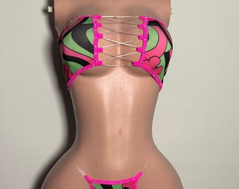 Può includere: Un set bikini a due pezzi con un vivace motivo ondulato rosa, verde lime e nero. Il top a fascia presenta un'allacciatura centrale con sottili cordini argentati, mentre lo slip a perizoma abbinato ha laccetti laterali trasparenti.