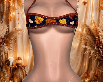 Può includere: Un set bikini a due pezzi in tessuto blu navy scuro con un motivo ripetuto di foglie autunnali e mele. Il top è in stile allacciato al collo e lo slip è a perizoma, entrambi rifiniti con tessuto color ruggine e piccoli strass trasparenti.