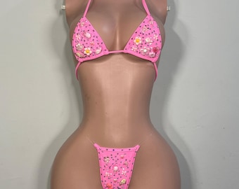 Può includere: Un set bikini rosa acceso con coppe a triangolo e slip a perizoma. Il tessuto è impreziosito da piccoli ciondoli colorati a forma di fiori e personaggi, insieme a strass trasparenti sparsi su tutto il capo.