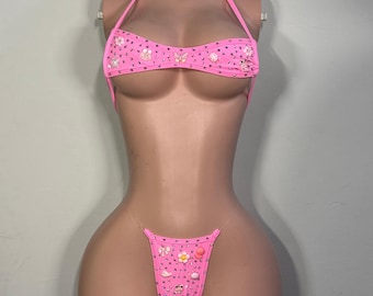 May include: A bright pink bikini set featuring a halter-style top and a thong-style bottom. Both pieces are embellished with numerous small clear rhinestones and various colorful charms, including flowers, butterflies, and small animal figures.