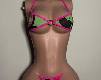 Può includere: Un set bikini a due pezzi con una vivace stampa grafica ondulata verde e nera con piccoli dettagli a forma di cuore. Il top ha un'allacciatura al collo, bordi rosa brillante e un anello centrale argentato a forma di cuore.