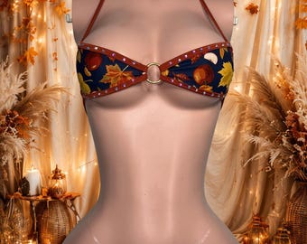 Può includere: Un set bikini a due pezzi in tessuto blu scuro con un motivo di mele rosse e foglie autunnali gialle. Il top presenta un'allacciatura al collo, un anello dorato centrale e bordi marroni con piccoli cristalli. Lo slip ha laccetti trasparenti.