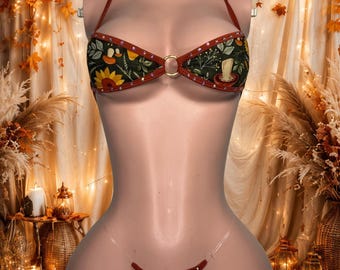 Può includere: Un bikini a due pezzi in tessuto verde scuro con stampa di funghi, girasoli e fogliame. Il top presenta un'allacciatura al collo, un anello dorato al centro e un bordo color ruggine con piccoli strass. Lo slip è dotato di laccetti trasparenti.