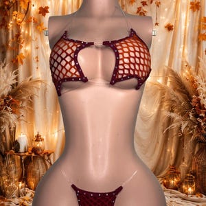 May include: A two-piece lingerie set featuring a dark red fishnet fabric with small silver-toned studs along the edges. The top consists of two separate cups connected by clear plastic straps, and the bottom is a matching thong with clear straps.