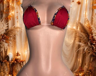 Può includere: Un set bikini a due pezzi con coppe triangolari rosso scuro e uno slip abbinato. I bordi delle coppe e dello slip sono rifiniti con un tessuto blu scuro decorato con un motivo a foglie dorate e bianche e piccole borchie metalliche.