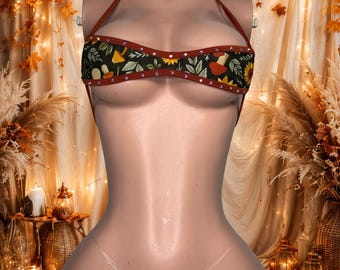 Può includere: Un set bikini a due pezzi in tessuto verde scuro con un motivo ripetuto di funghi arancioni, girasoli gialli e foglie verdi. Il top e lo slip sono bordati con tessuto color ruggine e decorati con piccoli strass trasparenti.