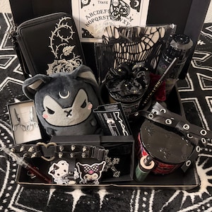 May include: A black gift box containing a dark gray plush rabbit, a black wallet with white rose embroidery, a black choker with silver spikes and a heart, a MAC mascara tube, a black scrunchie, a small glass bottle, and various stickers.