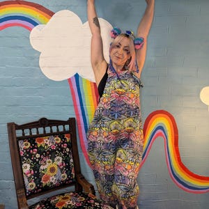 May include: A person wearing a colourful patterned jumpsuit against a rainbow backdrop. The person is wearing a headband with pom-poms.