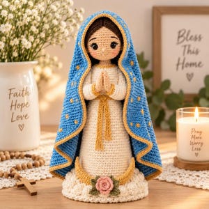 Puede incluir: Una muñeca de ganchillo de la Virgen María con un vestido color crema y una capa azul con capucha bordada con estrellas doradas. La figura se alza sobre una base de nube blanca con una rosa rosa, con las manos juntas en oración.