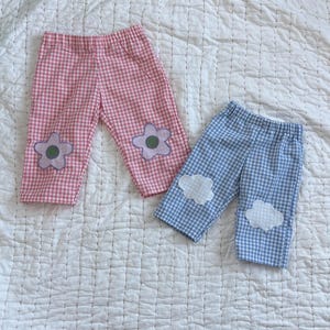 May include: Two pairs of pants with elastic waistbands. One pair features a pink and white gingham pattern with two light purple flower appliqués on the knees. The other pair features a blue and white gingham pattern with white cloud appliqués.