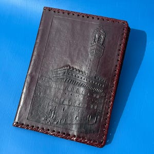 May include: A dark brown leather notebook cover featuring an embossed architectural design of a tall tower and a large building. The edges of the cover are finished with a decorative whipstitch lacing detail in a matching dark brown leather cord.