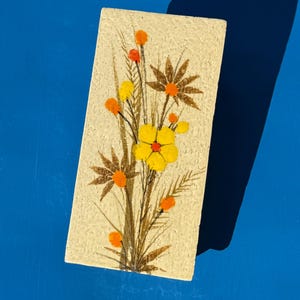 May include: A rectangular, cream-colored block with a textured surface featuring a painted floral arrangement. The design includes a central yellow flower, brown leaf silhouettes, and small orange and yellow circular accents on thin stems.
