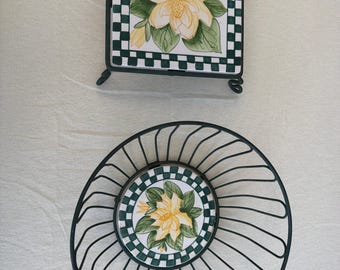 May include: A square ceramic tile trivet with a green and white checkered border and a yellow magnolia flower design, paired with a round black wire basket featuring a matching ceramic tile base with the same floral pattern.