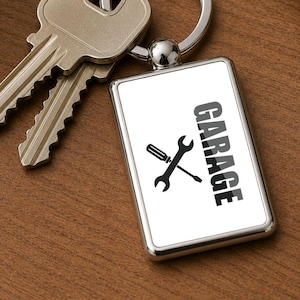May include: A rectangular metal keychain with a white insert featuring the black text GARAGE and a graphic of a crossed screwdriver and wrench. The keychain is attached to a silver split ring holding two standard brass-colored metal keys.