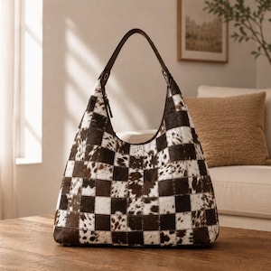 May include: A hobo-style handbag featuring a checkered pattern made of alternating squares of dark brown leather and white and brown cowhide fur. The bag has a single dark brown leather shoulder strap attached with metal hardware at the top.