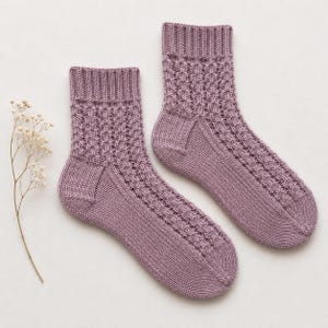 May include: A pair of mauve knitted socks featuring a ribbed cuff and a textured vertical pattern along the leg and top of the foot, with a smooth knit heel and toe section. A single dried sprig of baby's breath is positioned to the left.