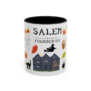 May include: A white ceramic mug with a black interior featuring the text SALEM FOUNDED 1626. The design includes a dark house, a flying witch silhouette, skeletons with pumpkin heads, autumn leaves, a black cat, a cauldron, and a coffee drink.