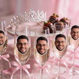 Peut inclure: Flûtes à champagne en verre transparent, chacune ornée d'un autocollant circulaire représentant une personne souriante avec des lunettes. Chaque flûte est décorée d'un ruban rose pâle noué autour du pied. En arrière-plan, un diadème et des fraises.