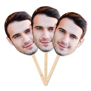 Può includere: Tre ritagli identici del volto di una persona con capelli corti castano scuro e barba leggera, ciascuno montato su un sottile bastoncino di legno chiaro. I ritagli presentano un'espressione sorridente con occhi marroni a ventaglio.