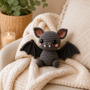May include: A dark gray crocheted bat plush toy featuring large black safety eyes, two small white fangs, pink blush accents on the cheeks, and inner ears lined with light pink yarn. The bat has outstretched wings and a rounded body.