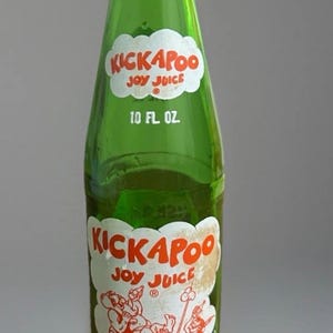 May include: A green glass bottle of Kickapoo Joy Juice soda with a white cloud-shaped label featuring red text and a cartoon illustration of characters in a wooden barrel. The bottle is marked with 10 FL OZ and the text MONEY BACK BOTTLE.
