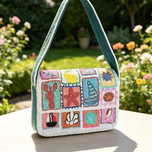 Peut inclure: Un sac à bandoulière rectangulaire doté d'une base blanche recouverte de patchs perlés colorés. Les patchs présentent divers motifs nautiques et côtiers, notamment un homard, une étoile de mer, un voilier, un crabe, un citron et des coquillages.