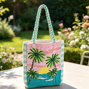 May include: A rectangular tote bag featuring intricate beadwork of a tropical sunset scene with palm trees, a yellow sun, and white boats on a turquoise sea. The bag has braided handles and side panels with alternating white and light green stripes.