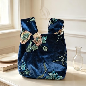 Könnte beinhalten: Eine tiefblaue Samthandtasche mit einem knotenförmigen Griff und aufwendigen Blumenstickereien. Das Design ist mit schimmernden Pailletten in Gold-, Türkis- und Grüntönen verziert, die ein strukturiertes Muster auf der Vorderseite erzeugen.