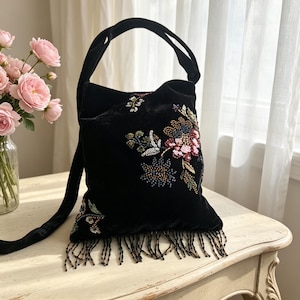 May include: A black velvet handbag featuring intricate floral embroidery with colorful beads and sequins. The bag has a single shoulder strap and a bottom edge adorned with long, dangling black beaded fringe, creating a textured appearance.