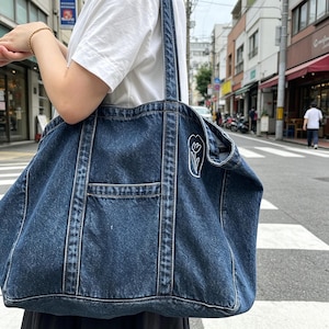 May include: A large dark blue denim tote bag featuring a white embroidered tulip outline on the side, two long shoulder straps, and a central exterior pocket with visible stitching details along the seams and edges of the fabric.