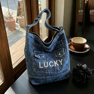May include: A blue acid-wash denim hobo bag featuring a single shoulder strap, two front pockets with silver-tone button closures, and the word LUCKY embroidered in white capital letters across the front panel of the bag.