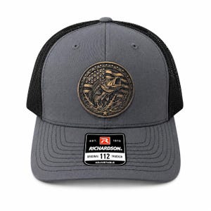 May include: A charcoal grey trucker hat with black mesh back panels and a circular bronze-toned patch featuring a bass fish jumping in front of an American flag. A black rectangular sticker on the brim reads EST. 1970 RICHARDSON 112 ORIGINAL ADJUSTABLE.