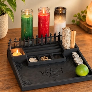 May include: A black rectangular tray featuring a fence-like border, a central pentagram and moon phase engraving, and multiple compartments. It holds three tall glass candles in green, red, and white, a sage bundle, palo santo, coins, jewelry, a watch, and a lime.