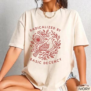 Pode incluir: Uma t-shirt de manga curta na cor marfim com uma estampa gráfica vermelha de um pássaro rodeado por vinhas florais. O texto "RADICALIZED BY BASIC DECENCY" está impresso em vermelho numa disposição circular em torno da ilustração.