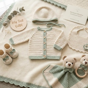 May include: A crochet baby set in cream and sage green, including a button-up cardigan, bonnet, booties, bib with a flower, a security blanket, and a bear rattle. A wooden disc reads My First Outfit and a card reads Heirloom Meadow.
