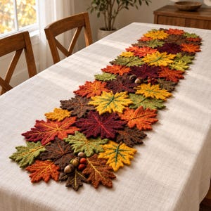May include: A table runner composed of numerous crocheted autumn leaves in shades of orange, yellow, red, brown, and green. The leaves are arranged in a dense, overlapping pattern, accented with small felted acorns scattered throughout.