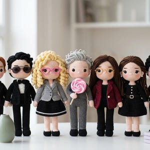 May include: A collection of seven crocheted dolls standing in a row. The dolls feature various outfits including black suits, a grey blazer with a black skirt, a grey suit holding a pink lollipop, and black dresses. Each doll has distinct hair.