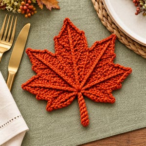 Peut inclure: Un dessous de verre au crochet orange brûlé en forme de feuille d'érable. Le dessous de verre présente un motif de mailles tricotées à la main et texturées, avec des nervures en relief partant du centre vers les lobes pointus.