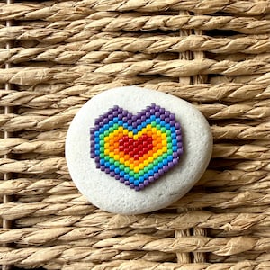 Peut inclure: Une pierre ovale blanche et lisse ornée d'un motif en forme de cœur composé de petites perles multicolores. Le perlage présente des couches concentriques de violet, bleu, vert, jaune, orange et rouge, formant un motif arc-en-ciel vibrant.